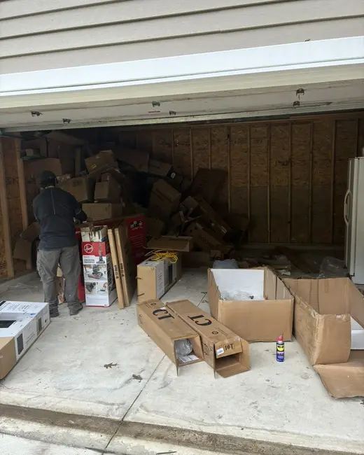 Garage filled with cardboard boxes, packaging and household items, with a person sorting near the back wall