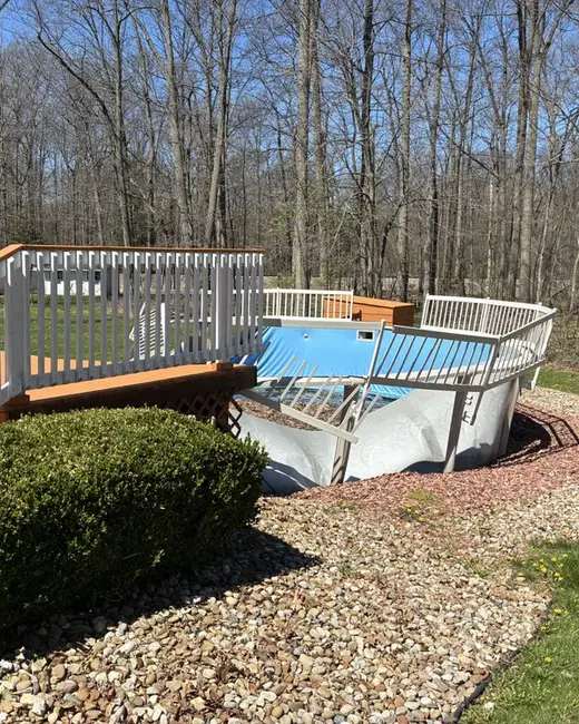 Above-ground pool with blue liner and white railing standing beside a wooden deck and shrub bed