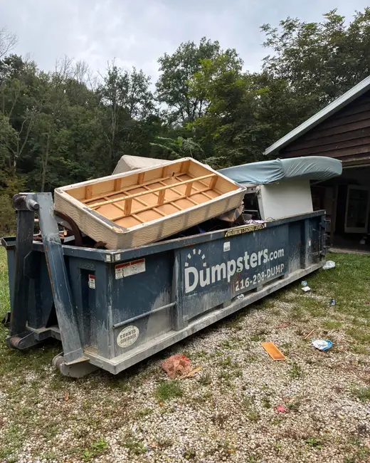 Roll-off dumpster filled with wooden furniture and debris, set on gravel beside a house